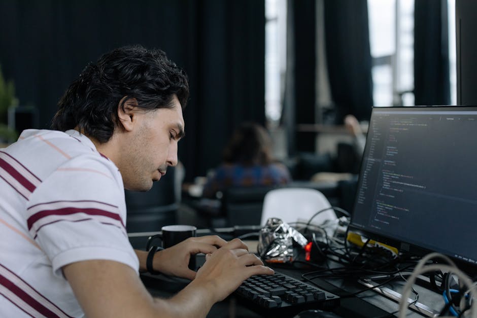 A young man with dark, wavy hair is focused on typing code on a computer keyboard in an indoor workspace. The desk in front of him has a large monitor displaying lines of programming code, along with 