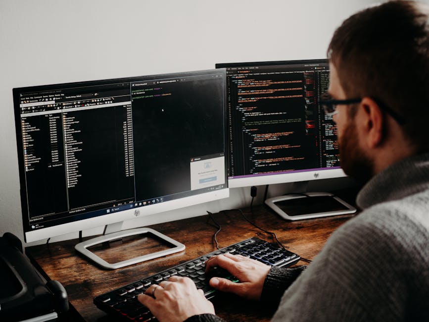 A young man with dark, wavy hair is focused on typing code on a computer keyboard in an indoor workspace. The desk in front of him has a large monitor displaying lines of programming code, along with 