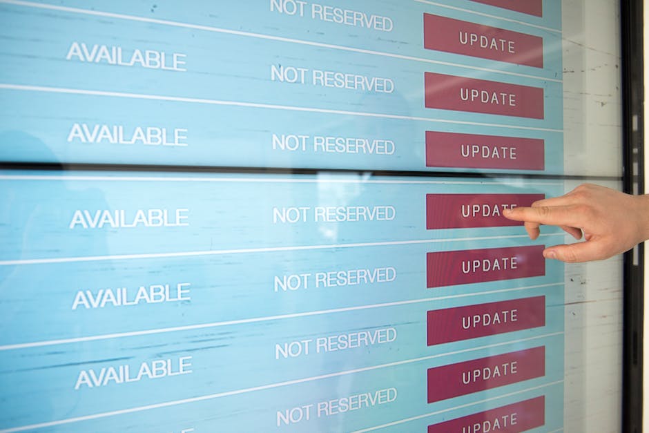 A person's hand with light-colored skin and short, clean nails is pressing the red 'UPDATE' button on a digital display board that shows multiple rows of information, including the words 'AVAILABLE' a